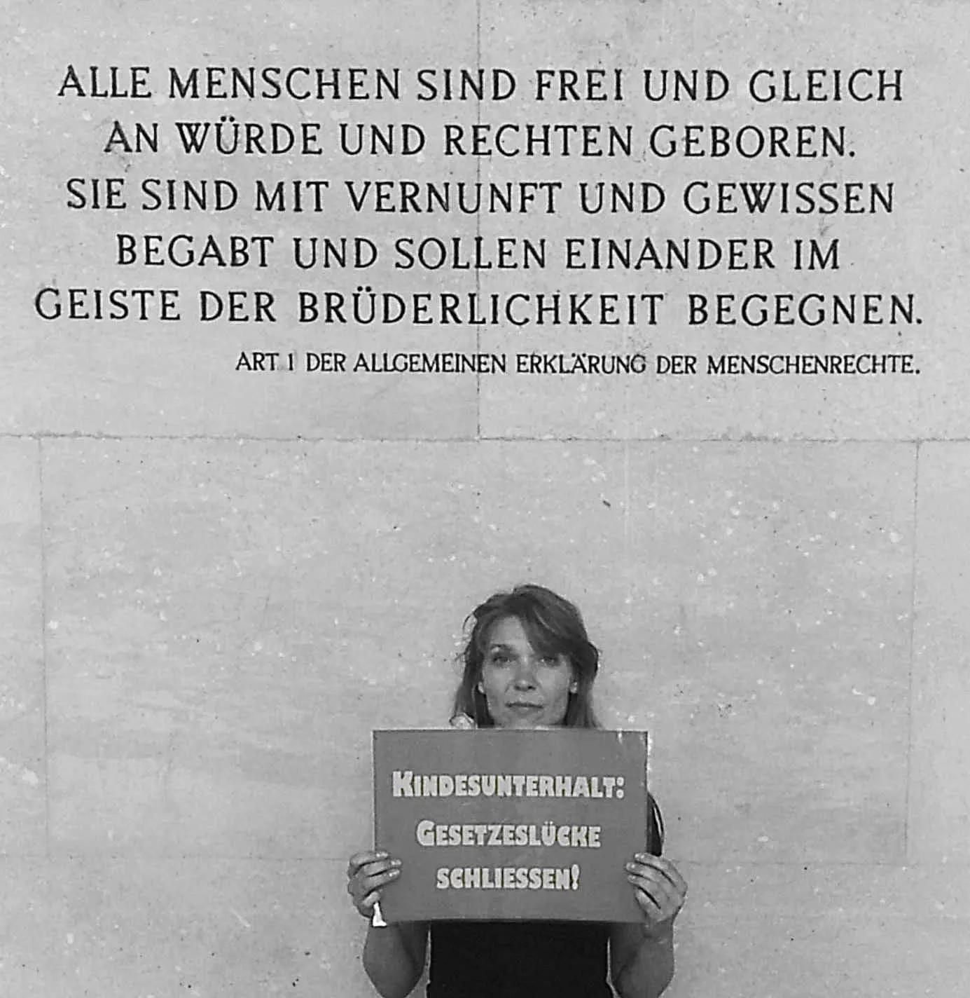 Maria Stern hält ihr Schild „Kindesunterhalt: Gesetzeslücke schließen!“ unter der Inschrift von Artikel 1 der Menschenrechtserklärung