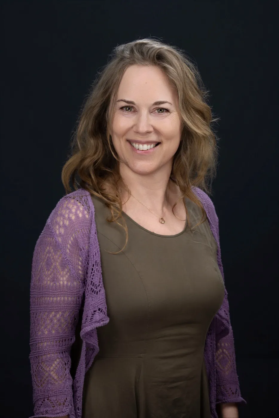 Angela Cooper mit langen blonden Locken in olivgrünem Kleid und fliederfarbener Strickjacke vor dunkelblauem Hintergrund